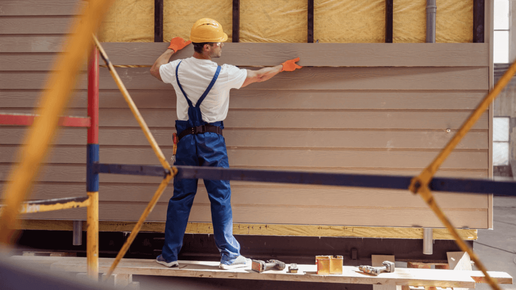 A man in overalls and a hard hat standing on a platform, doing a siding service