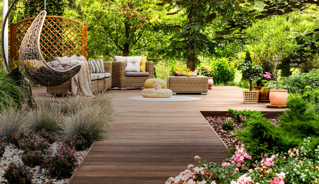 Backyard full of trees, flowers and bushes, with a wooden decking decorated with armchairs and a small table containing a jug of juice and fruits, transmitting a cozy atmosphere.