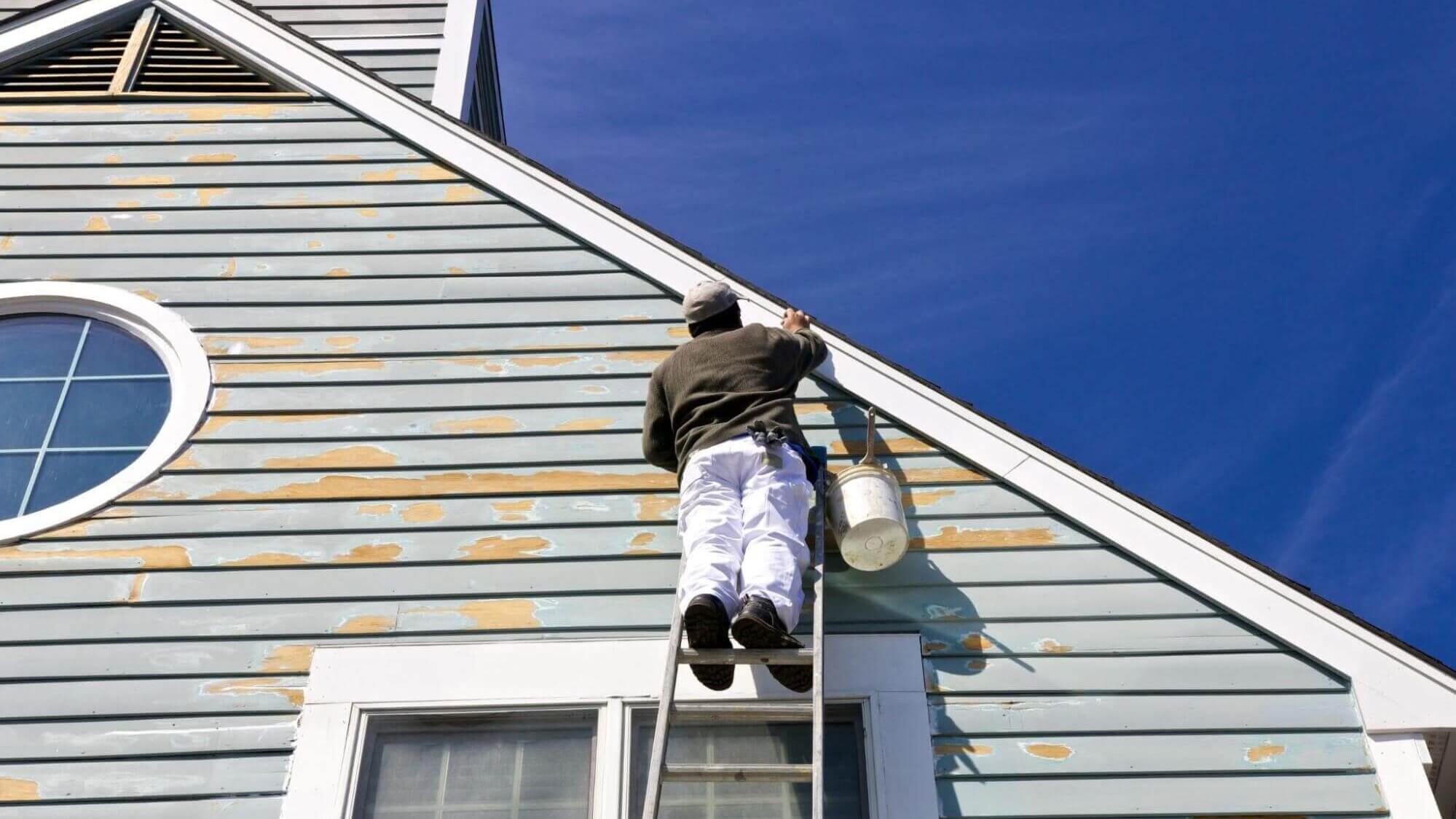 A man, on a ladder, enhancing the home's aesthetics with a trimming service, against a backdrop of a sunny day and clear blue sky.