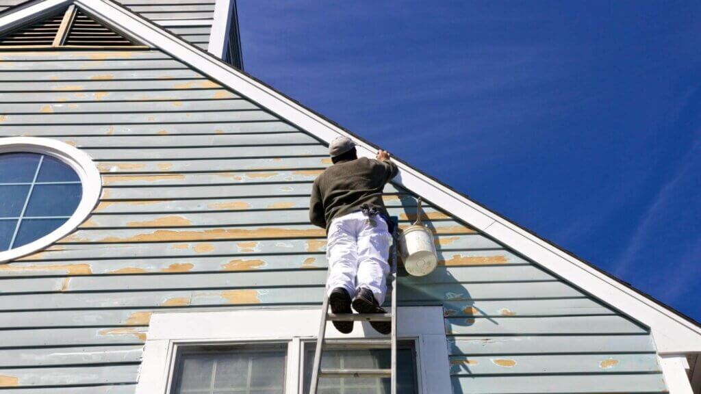 A man, on a ladder, enhancing the home's aesthetics with a trimming service, against a backdrop of a sunny day and clear blue sky.