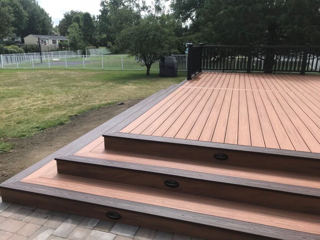 wood deck with a three steps stair, a fence in the back, trees in the background and grass on the left.